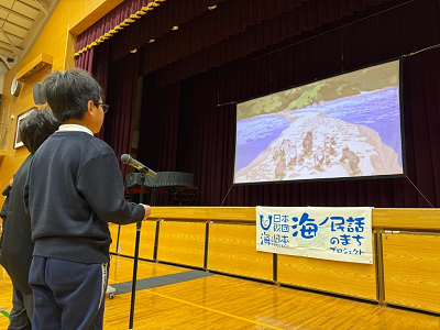 マイクの前に立った児童が壇上のスクリーンを見ている写真