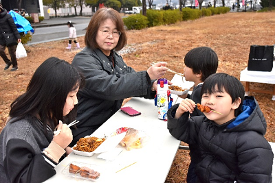 屋外に設置された机で焼きそばやから揚げを食べる子供たちの写真