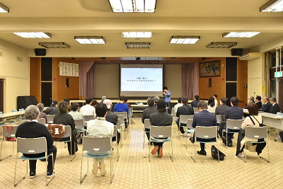 パイプ椅子に座った人たちにスクリーンを使って説明している人の写真