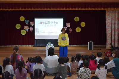 黄色のシャツに青いズボンを履いた女性が園児たちの前に立っている写真