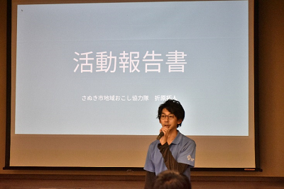 「活動報告書」と表示されているスクリーンの前でマイクを持っている男性の写真