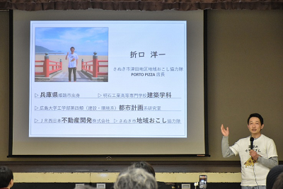 「折口洋一」のプロフィールが表示されているスクリーンの前でマイクを持っている男性の写真