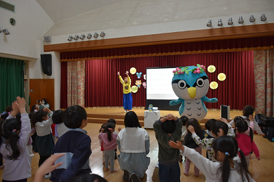 園児達と一緒に両手を上にあげる女性と鳥のキャラクターの写真