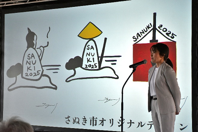 3種類のイラストが映し出されたスクリーンの前で話すスーツ姿の女性の写真