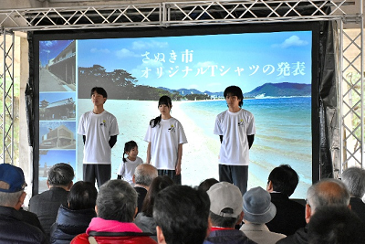 海の写真が映し出されたスクリーンをバックにTシャツを着た男女が立っている写真