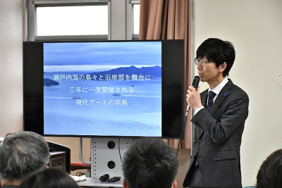 モニター横でマイクを手に話すスーツ姿の男性の写真