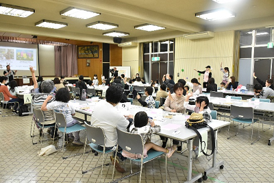 座っている親子たちが前方に立つ男性に向かって片手をあげている写真