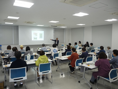 大勢の前でスクリーンを指しながら話す男性の写真