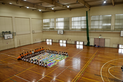 床に置かれた巨大な絵の周りに座る園児と大人達の写真