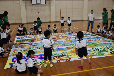 床に置かれた巨大な絵の周りで座ったり立っている園児達の写真