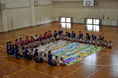 床に置かれた巨大な絵の周りに座っている園児や大人たちが手を振っている写真