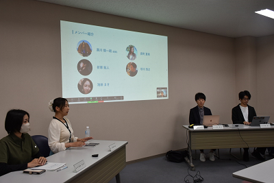 メンバーの紹介スライドが映っているスクリーンの前で着席している人たちの写真