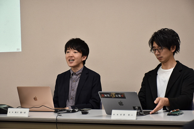 ネームプレートが置かれた机に着席している男性2人の写真