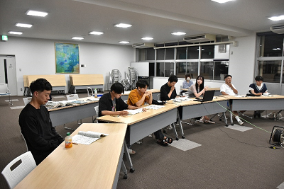 室内でアーチ型に並べた長机の座席に6名の男女がつき手元にある資料を読んでいる写真