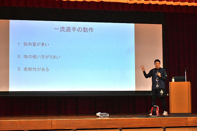 体育館の舞台に立って話す男性の写真