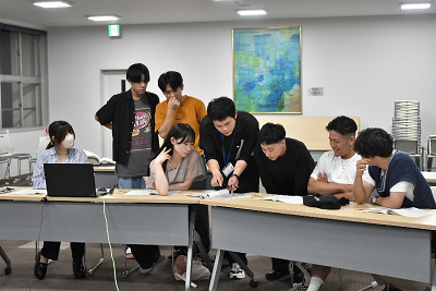 室内で8名の男女が長机に置かれた資料を読むために密集している写真