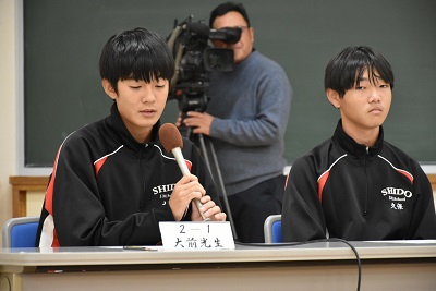 マイクを持った生徒の後ろにカメラを抱えた人が立っている写真