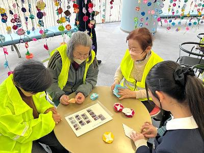 高校生にアサガオの折り方を教わる交通安全母の会のメンバー