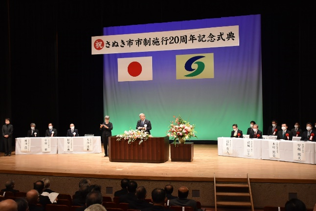 講堂で祝さぬき市市制施行20周年記念式典と書かれた横断幕の下日本国旗とさぬき市の市章旗が掲げられ、壇上に講壇の左右に白い長机が置かれ参列者が10名程度座るなか講壇に立っている男性がスピーチしているのを客席にいる人たちが聞いている写真