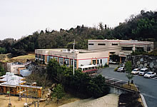 四角い建物の周りに駐車場や樹木が植わっている写真